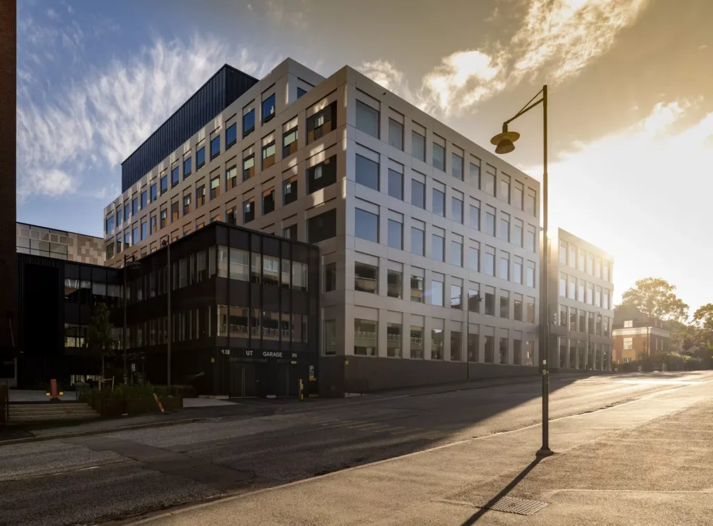 Byggnad med moderna glasfönster i motljus från solen, gatlyktor och trottoar i förgrunden.