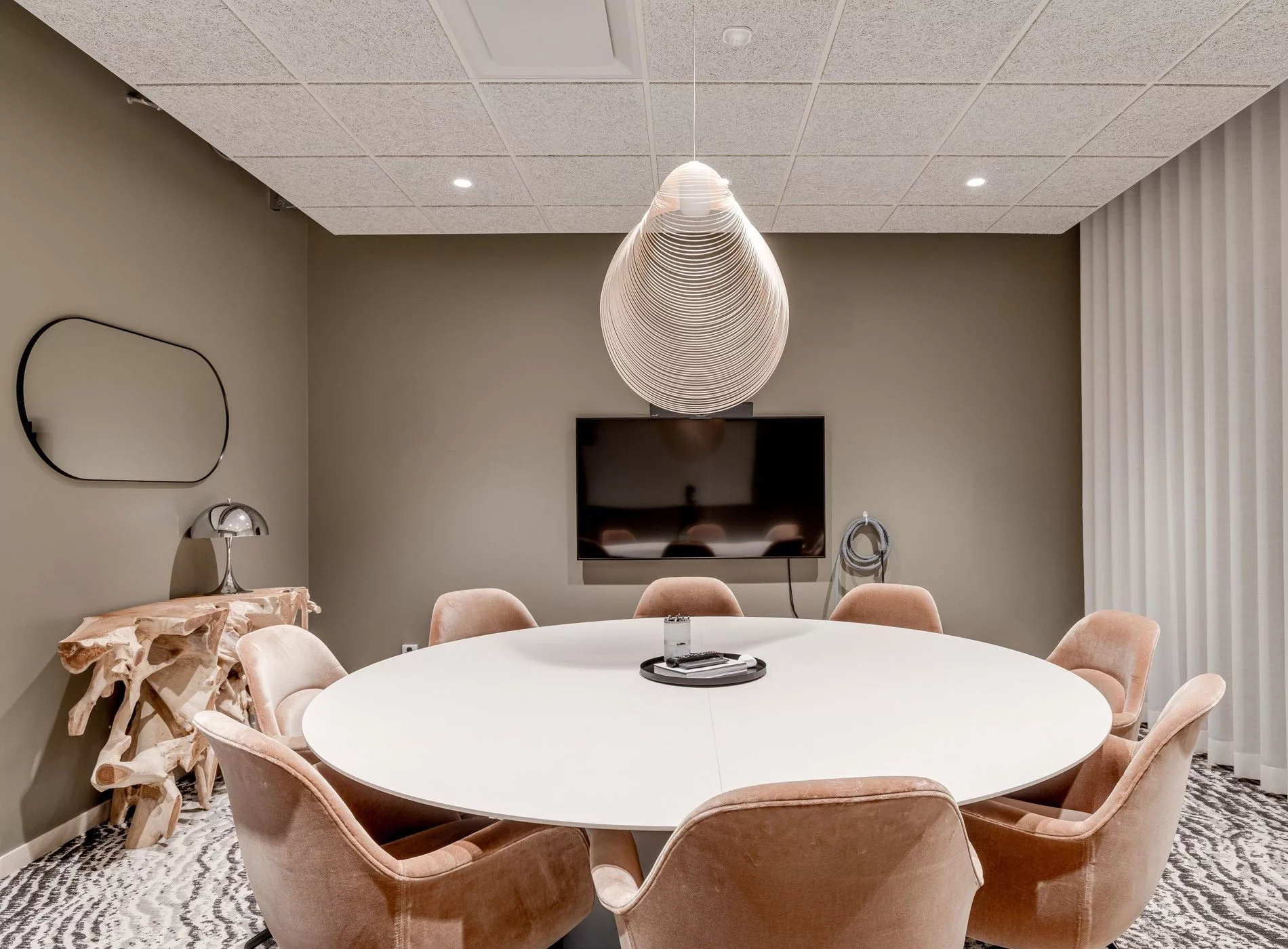 Ett mötesrum med ett runt vitt bord, beige stolar, en hängande lampa och en väggmonterad skärm.