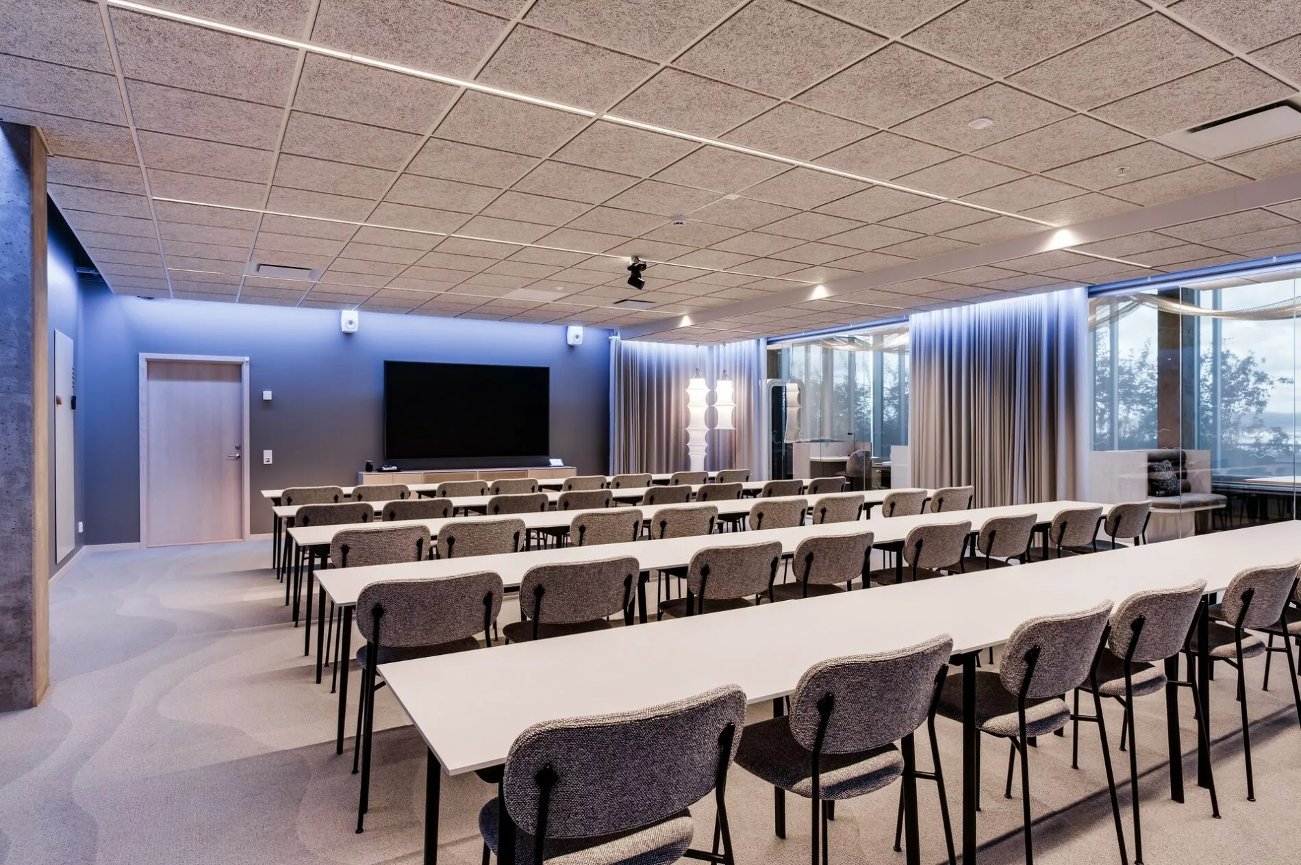 En konferenssal med flera rader av vita bord och grå stolar, samt en stor skärm längst fram.