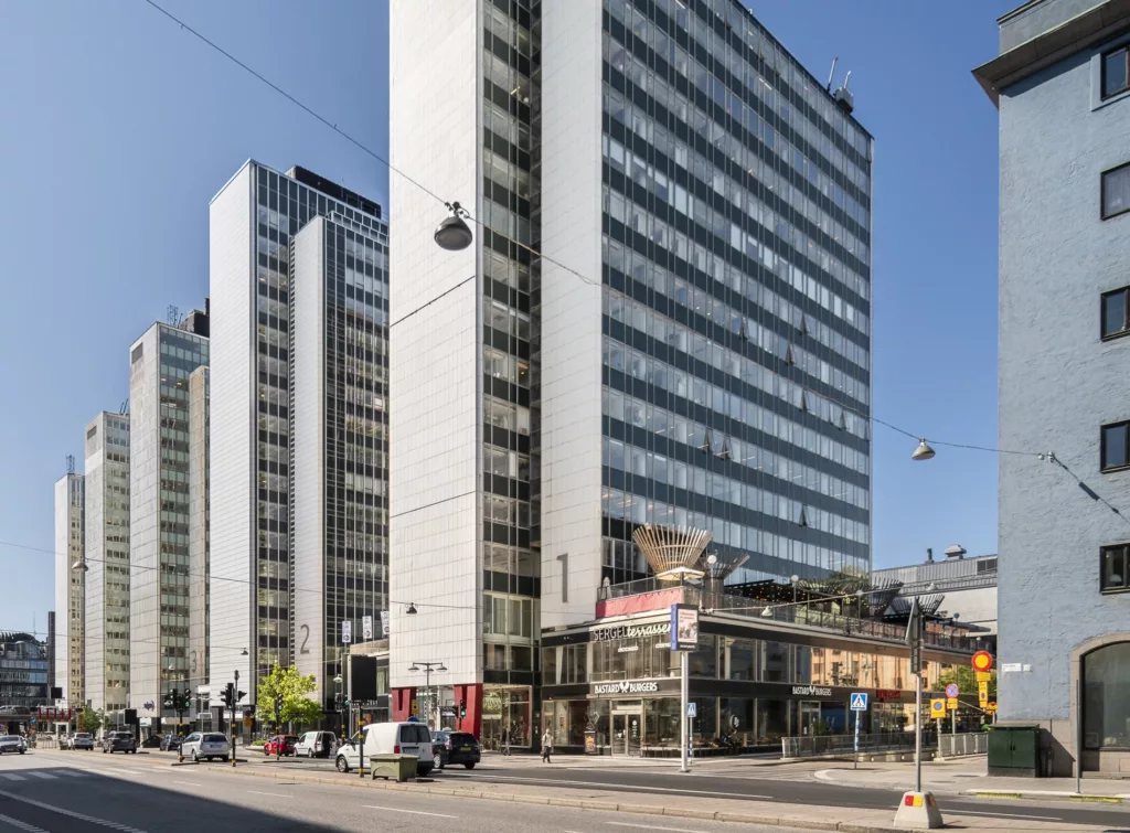 Höga kontorsbyggnader med glasfasader på en stadsgata, omgivna av bilar och fotgängare under en klarblå himmel.
