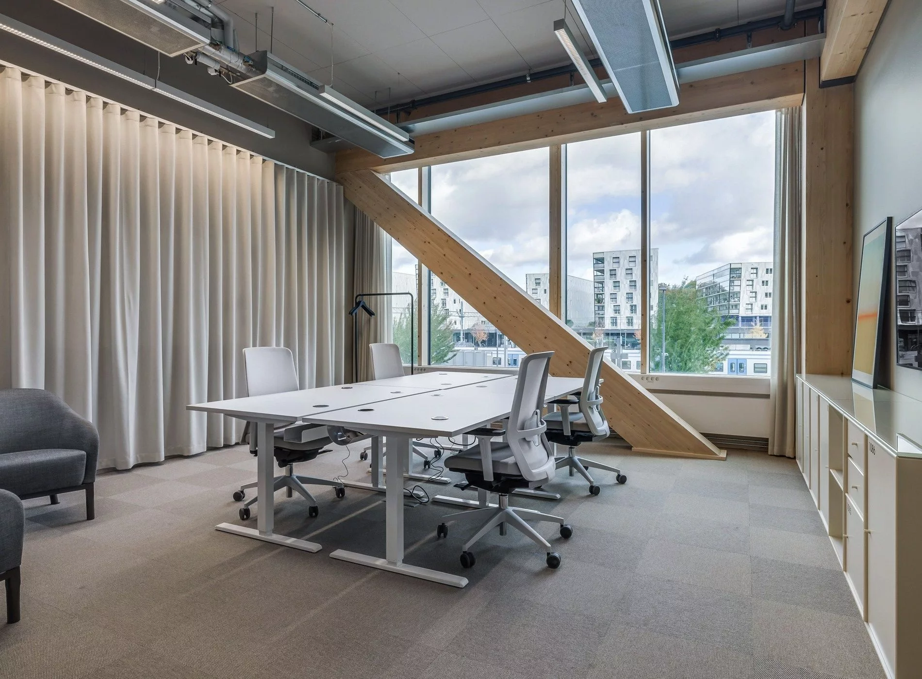 Ett kontorsrum med vita skrivbord och stolar, stora fönster och en synlig träbjälke som en del av arkitekturen.