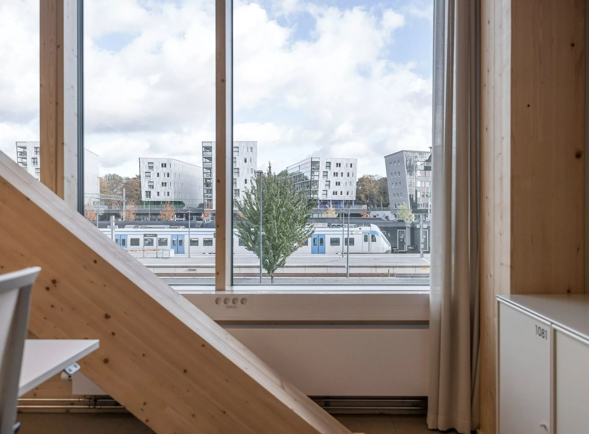 Utsikt genom ett stort fönster med ett träd och moderna byggnader utanför, samt en trappa i förgrunden.