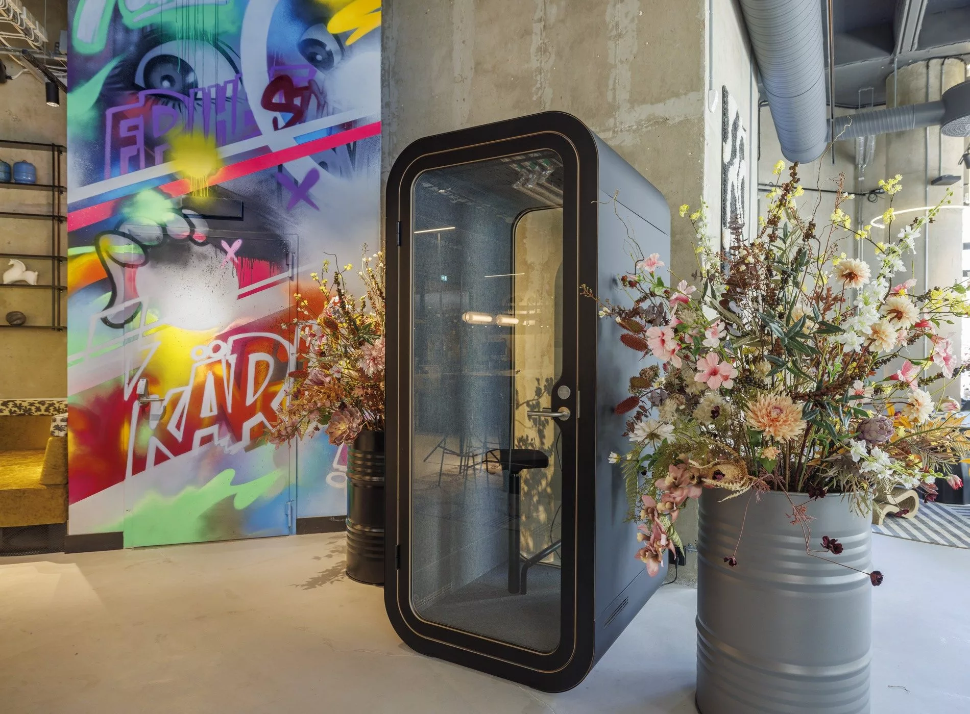 Liten ljudisolerad telefonkiosk omgiven av blomkrukor, med en färgglad graffitivägg i bakgrunden.