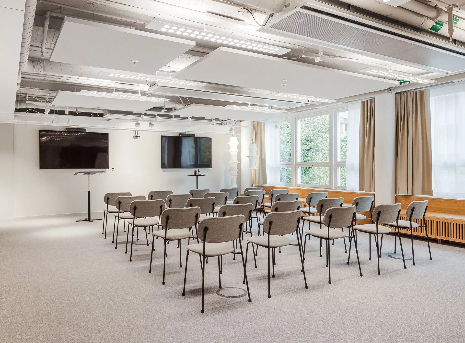 En konferenssal med stolar i rader, två stora skärmar och stora fönster med ljusinsläpp.