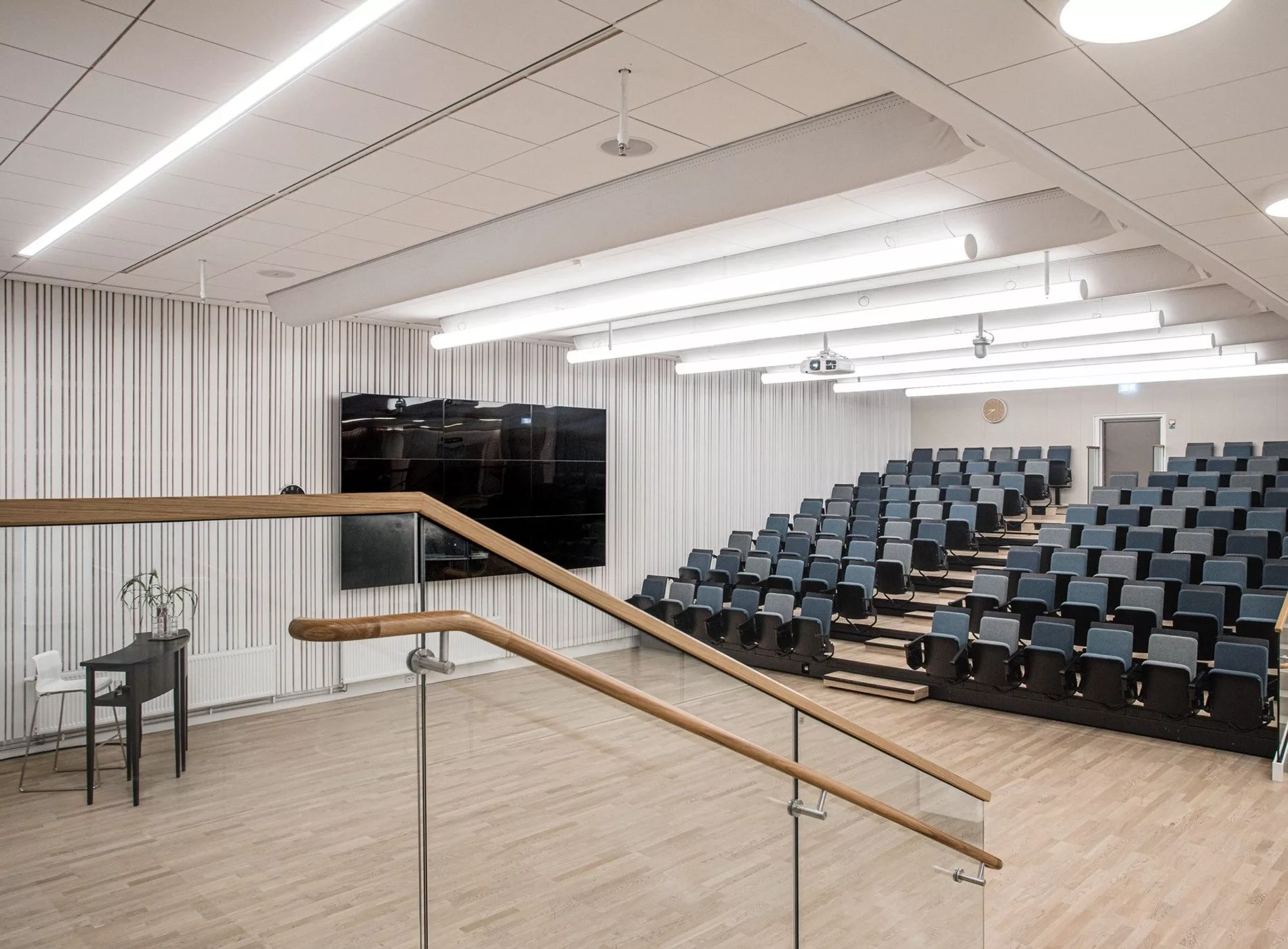 Bild på en föreläsningssal med gradängsittning, en stor väggmonterad skärm och en trätrappa med glasräcke.
