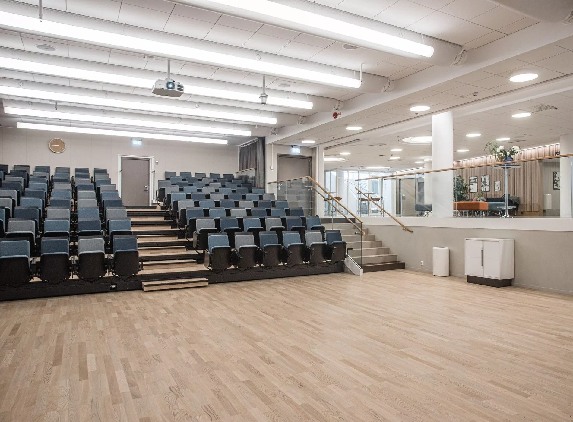 Bild på en föreläsningssal med gradängsittning, projektor i taket och en glasvägg som vetter mot en annan del av byggnaden.