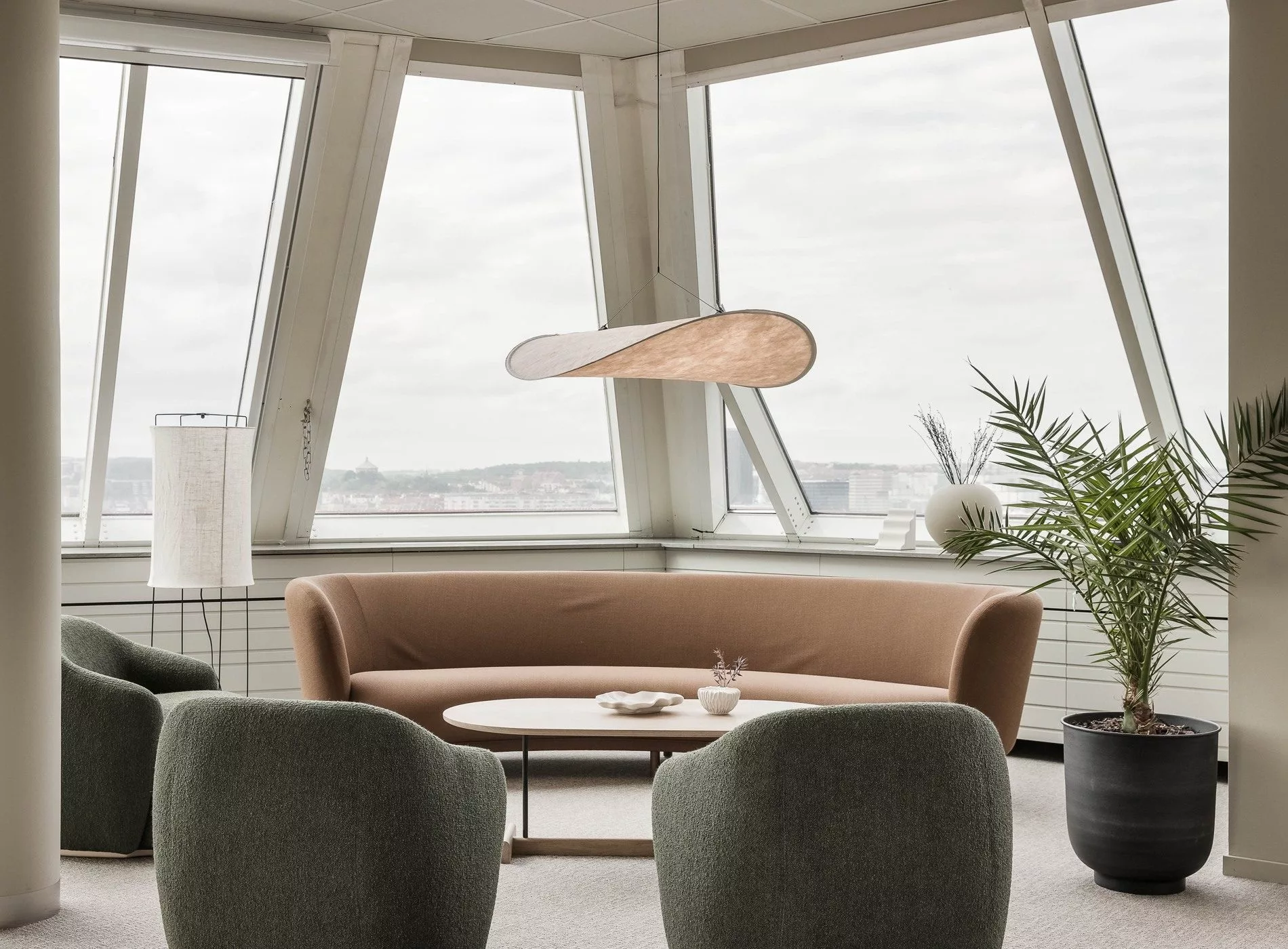 Ett modernt rum med panoramafönster, en beige soffa, gröna fåtöljer och en hängande lampa.