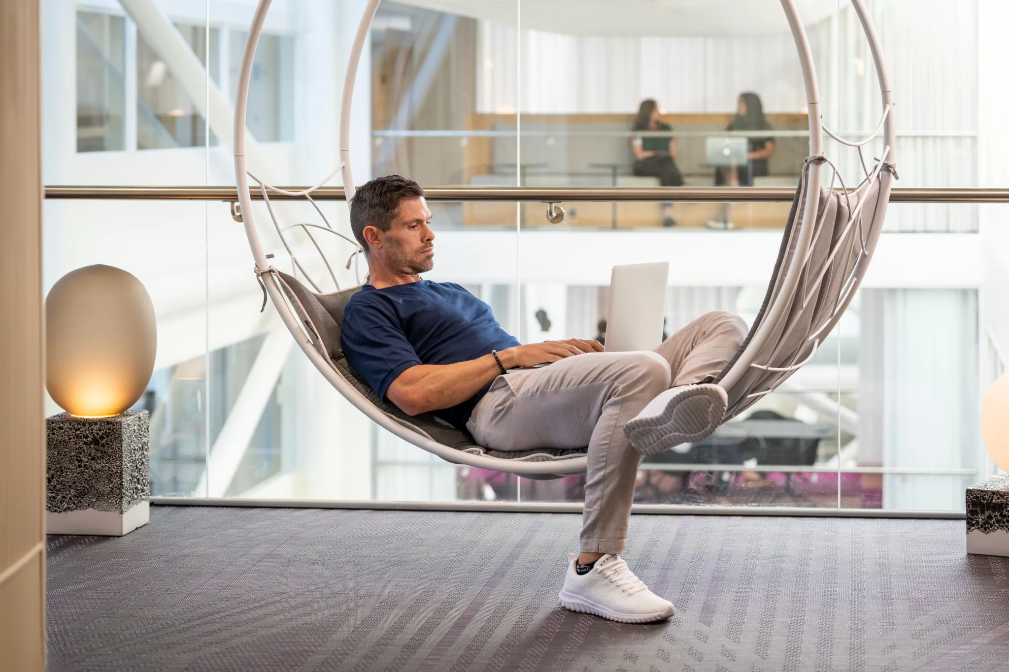 En man sitter i en hängande stol med en laptop i en modern kontorsmiljö med glasväggar.