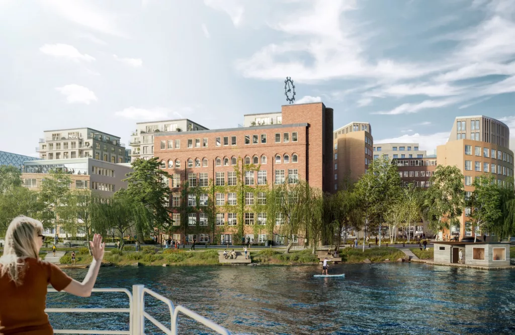 Vy av en stad med en stor tegelbyggnad, grönska och en flod i förgrunden. En person står på en brygga till vänster.