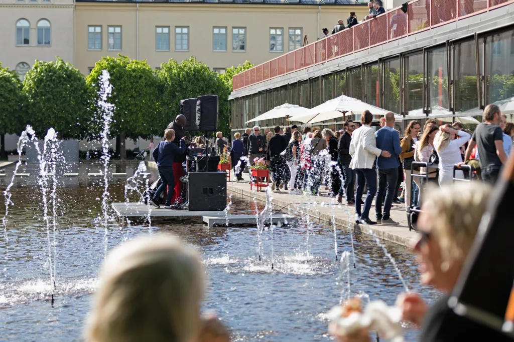 2. Fontän vid ett torg med människor, en musiker spelar på en scen.