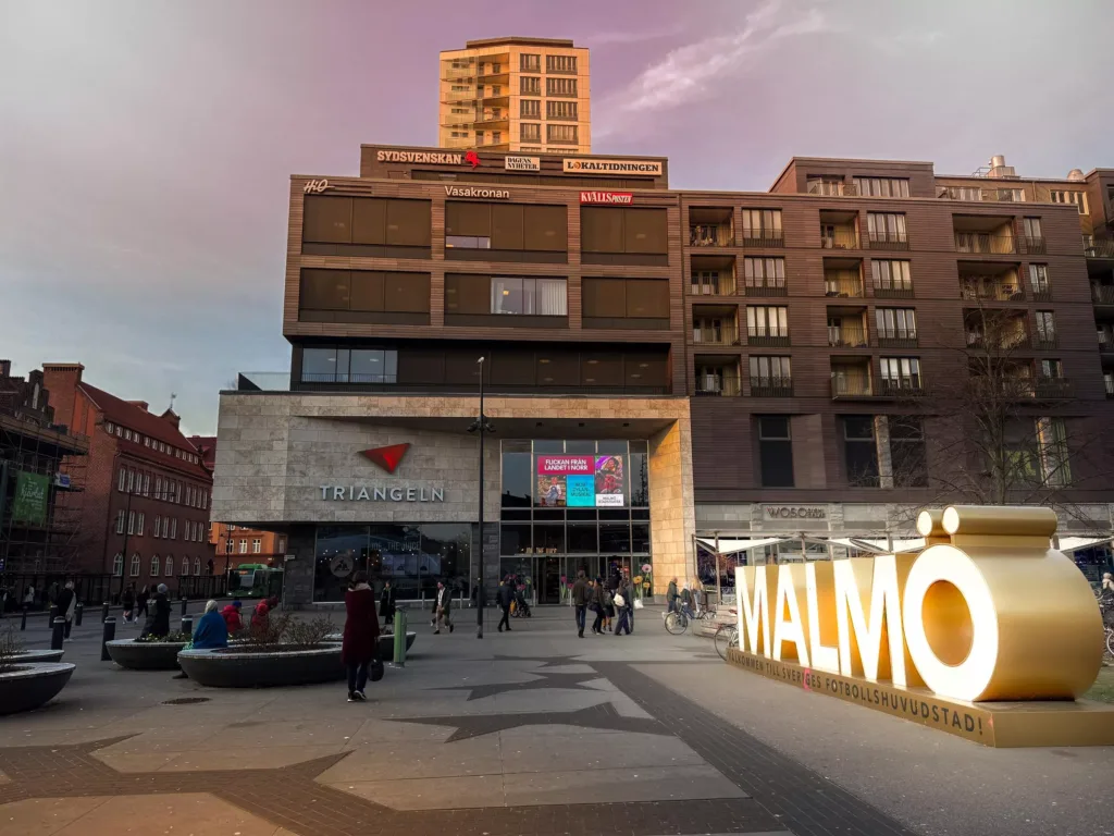En byggnad med skylten "Malmö" framför, omgiven av människor och stadsmiljö under en rosa kvällshimmel.