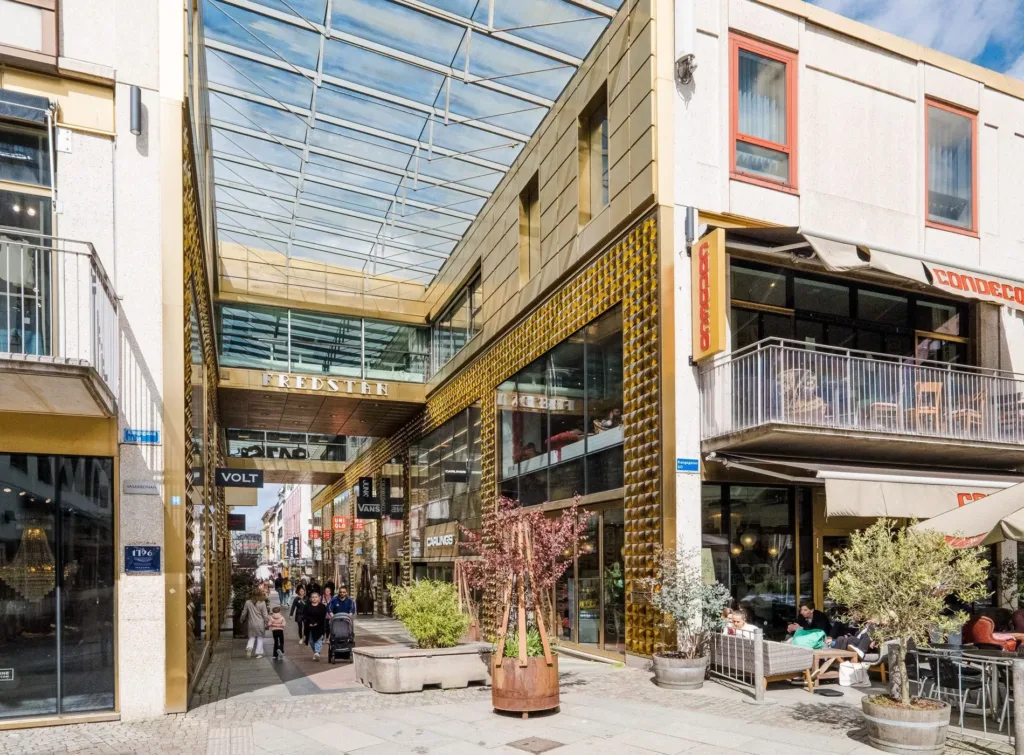 Ett shoppingområde med moderna byggnader, glasfasader och dekorationer, med människor som promenerar längs gångvägen.