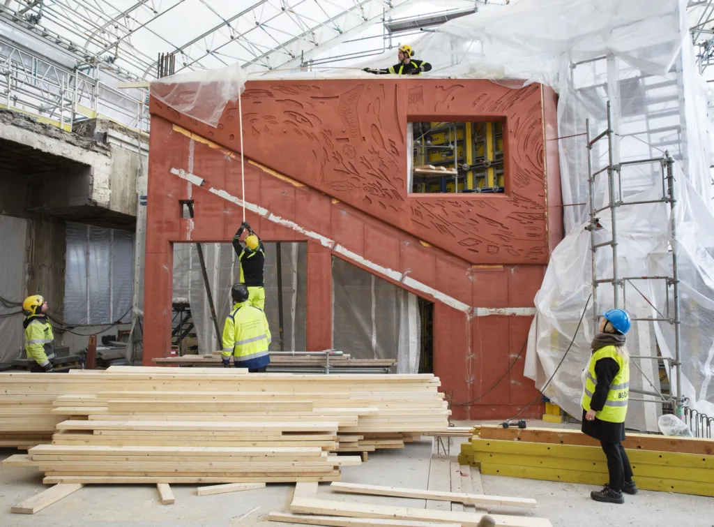 Ett orange byggnadselement under konstruktion, omgiven av byggnadsarbetare och trävirke.