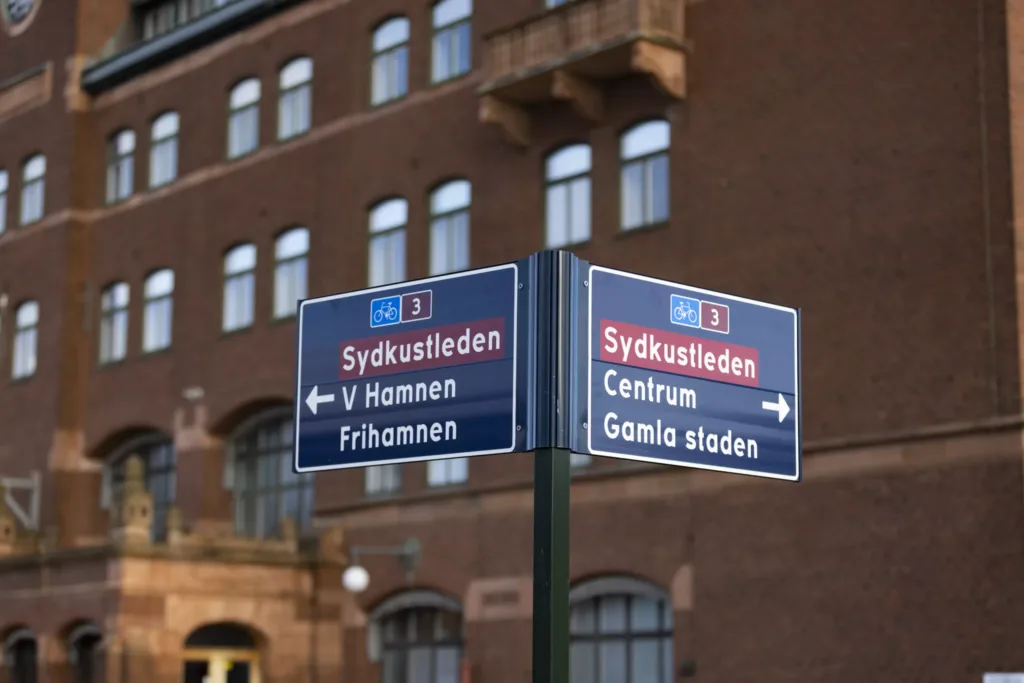 Vägskyltar med texten "Sydkustleden" som visar riktningar mot "V Hamnen, Frihamnen" och "Centrum, Gamla staden" med en tegelbyggnad i bakgrunden.