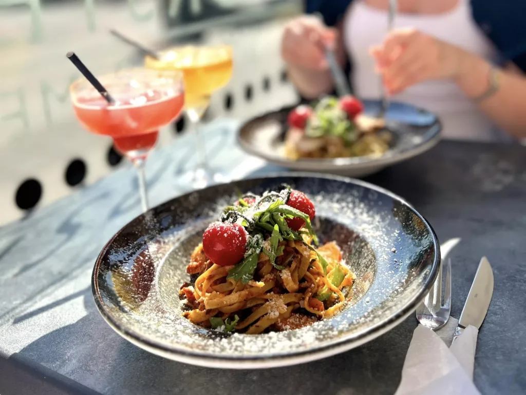 En tallrik med pasta toppad med körsbärstomater och sallad, med drinkar och en annan tallrik i bakgrunden.