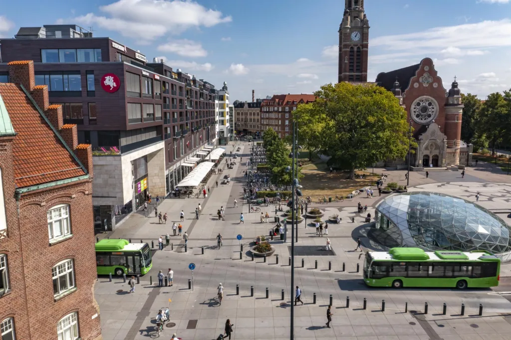 En stadstorg med människor, gröna bussar, en kyrka och moderna byggnader under en solig dag.