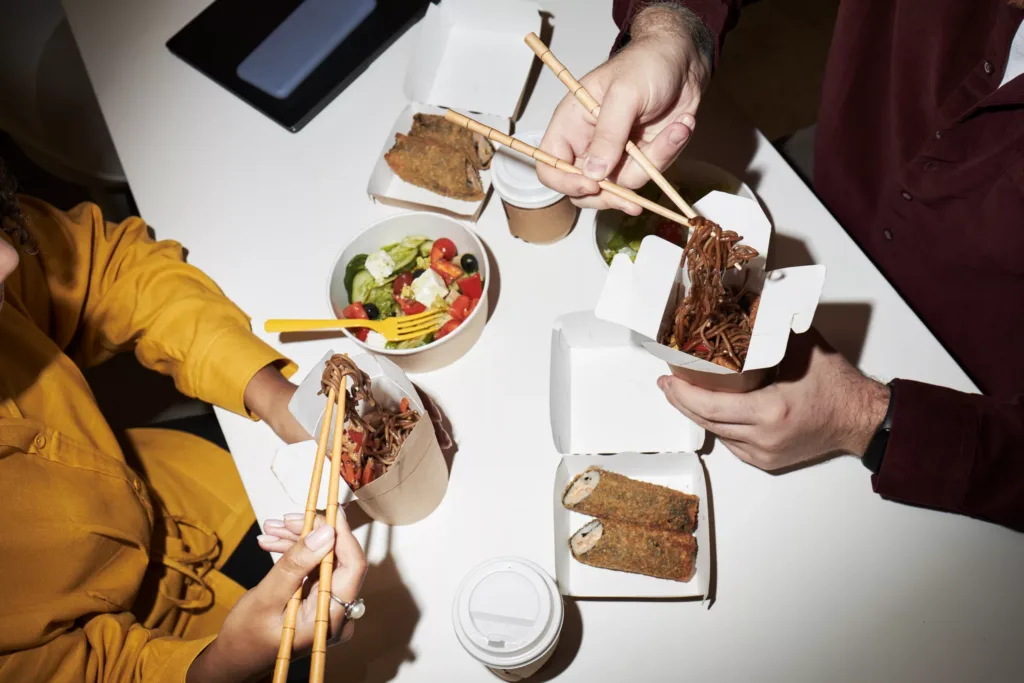 Caucasian man and Black woman eating Asian takeout noodles and spring rolls with chopsticks at table, sharing meal with coffee cups and salad, hands visible interacting with food