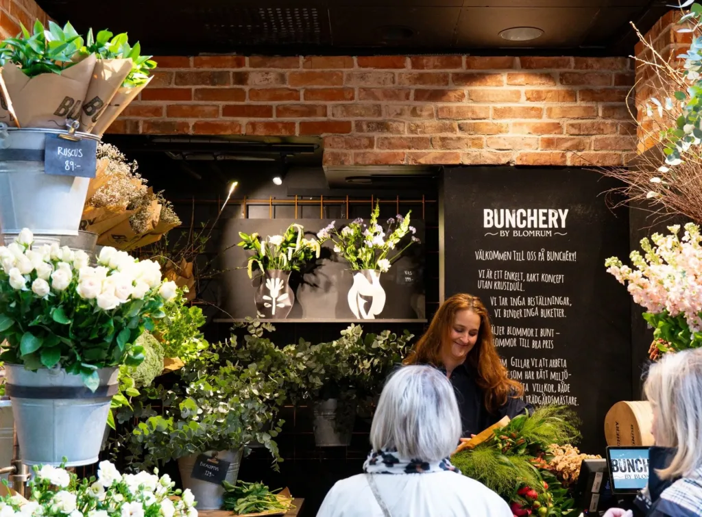 En kvinna arbetar med blommor i en blomsterbutik med tegelväggar och skyltar som visar butikens namn "Blomstery."