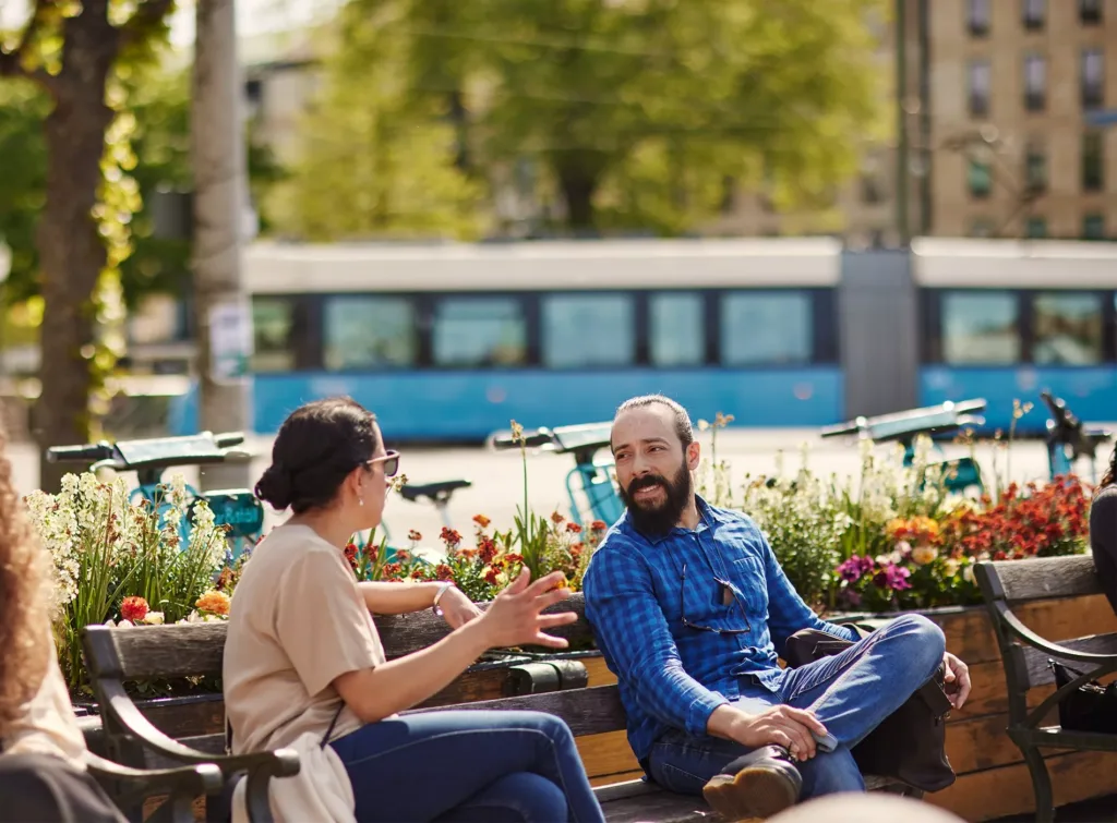 Två personer sitter utomhus vid en blomsterprydd bänk och pratar. En blå spårvagn syns i bakgrunden.