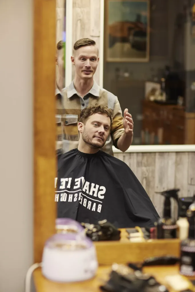 4. En frisör arbetar med en kund som sitter i stolen med en cape med texten "SHAVE IT" tryckt på.