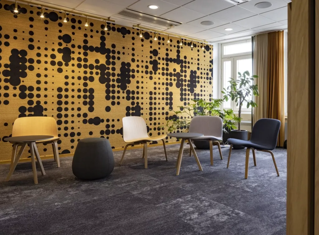 En lounge med trästolar, en svart pall och en dekorativ vägg med prickmönster, samt växter och stora fönster.