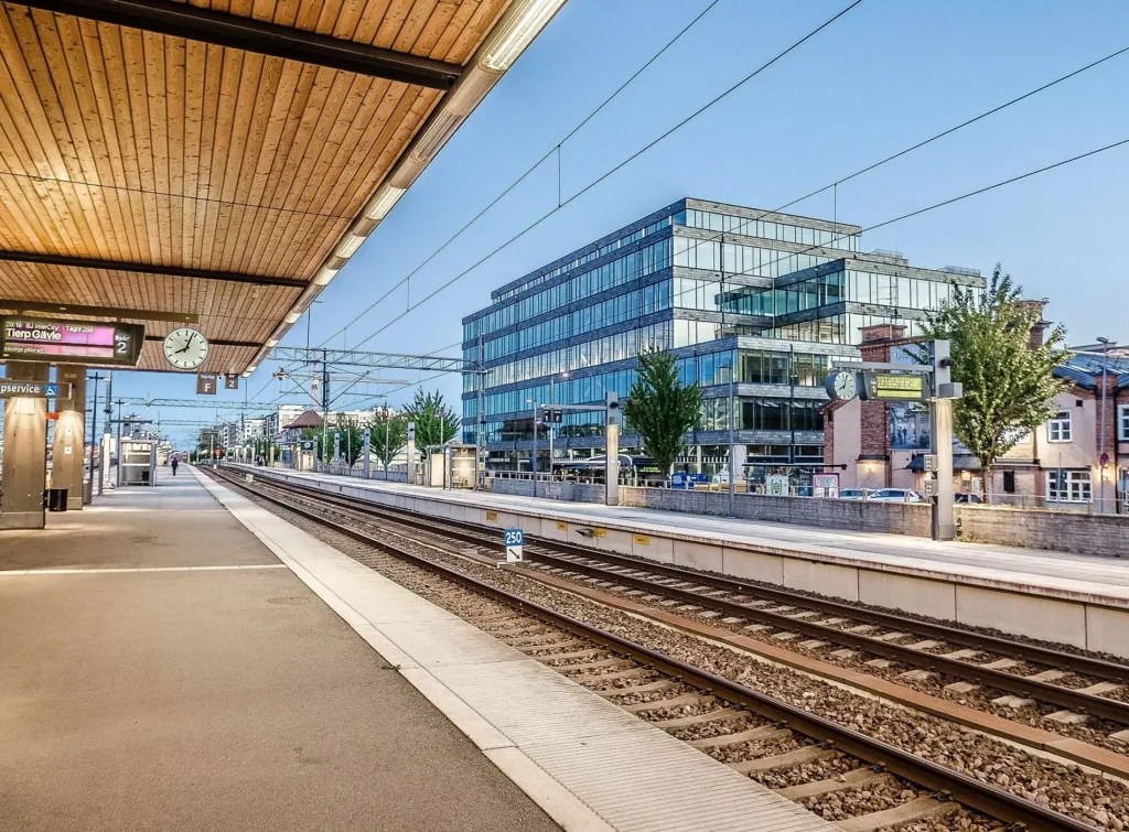 En modern tågstation med plattformar och spår, omgiven av glasfasader och byggnader under en klar himmel.