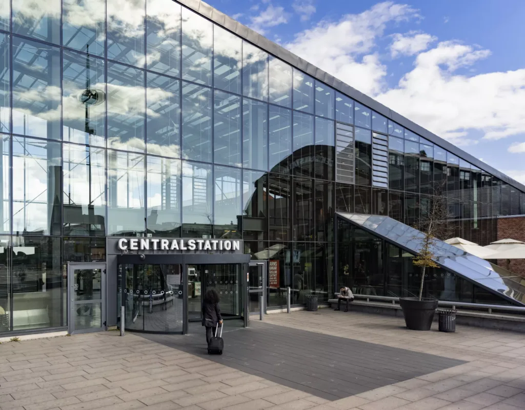 Centralstationens glasfasad med texten "Centralstation" vid ingången, omgiven av trottoar och blå himmel med moln.