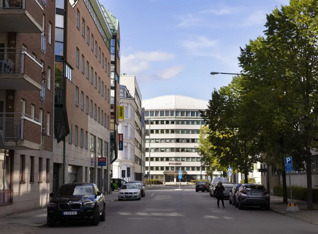 En stadsgata med parkerade bilar, träd och moderna kontorsbyggnader under en ljus blå himmel.
