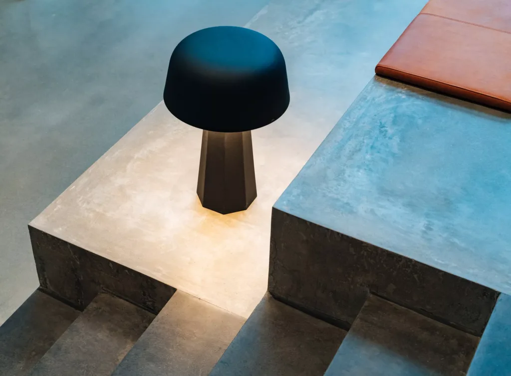 En modern bordslampa med mörk skärm placerad vid betongtrappor och en orange sittdyna.