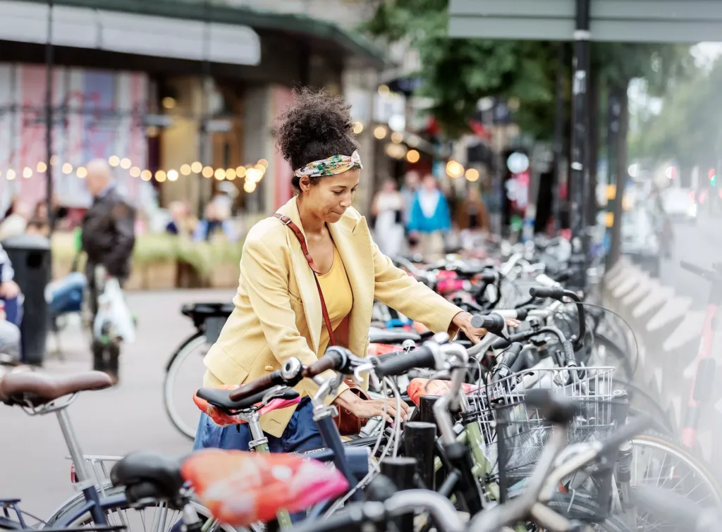 En kvinna i gul kavaj håller i en cykel bland många parkerade cyklar på en livlig gata.