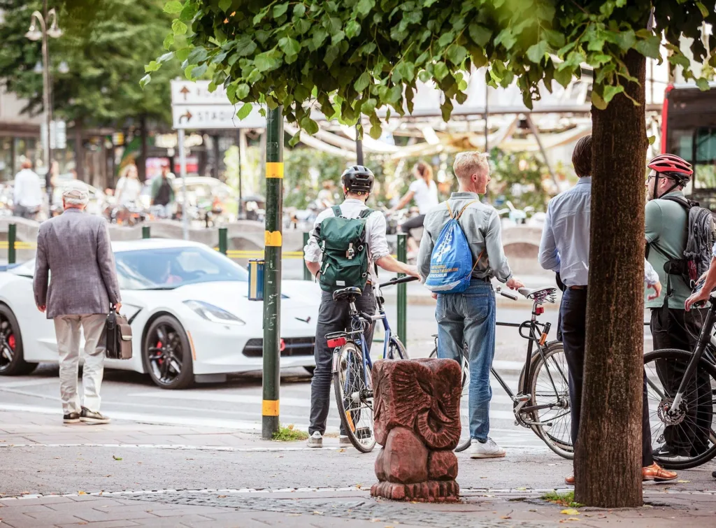 En stadsmiljö där flera personer med cyklar står nära en vit bil under träd.
