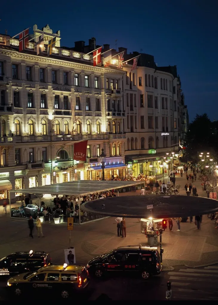 Kvällsvy över livlig stadsgata med upplysta byggnader och folk som promenerar.