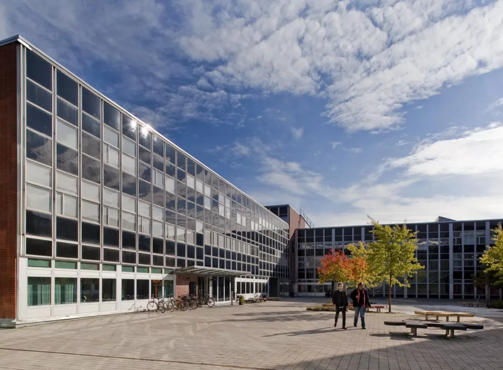 En rymlig innergård med moderna byggnader, träd och en klar blå himmel. Två personer står nära ett träd med höstfärger.