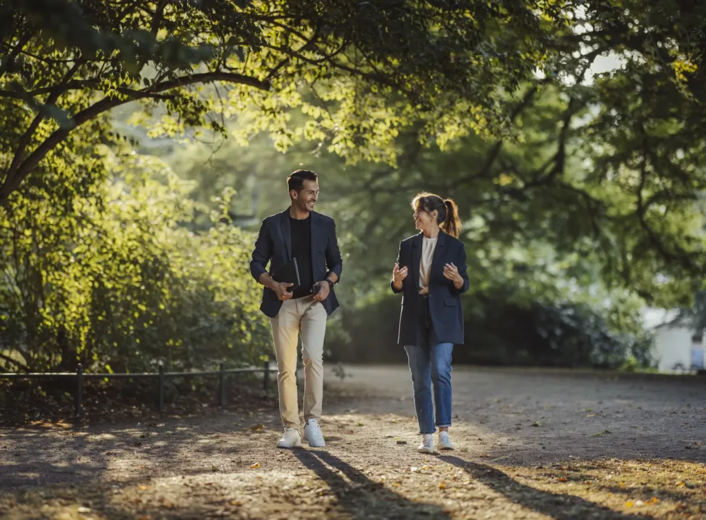 Två personer promenerar i en park under träden, engagerade i ett samtal.