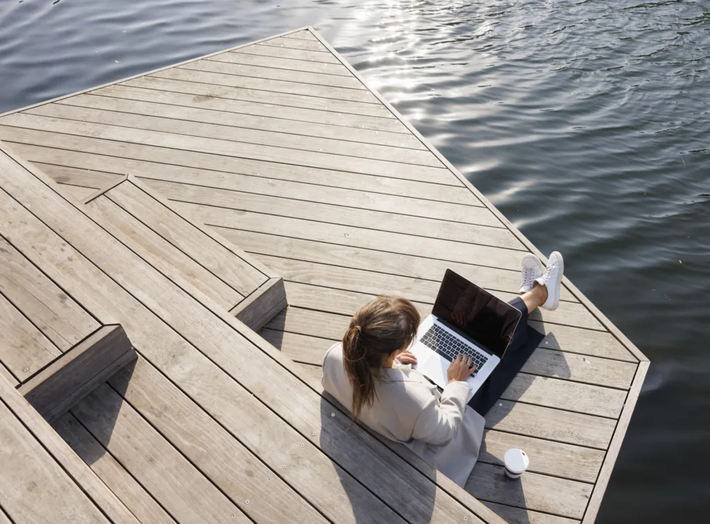 4. Person sitter på en träbrygga vid vattnet och arbetar på en laptop.