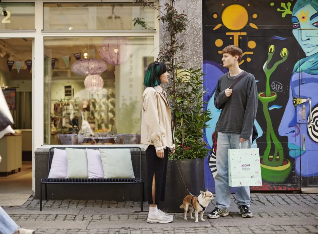 Två personer står och pratar utanför en butik med färgglad väggmålning, en liten hund vid deras fötter.