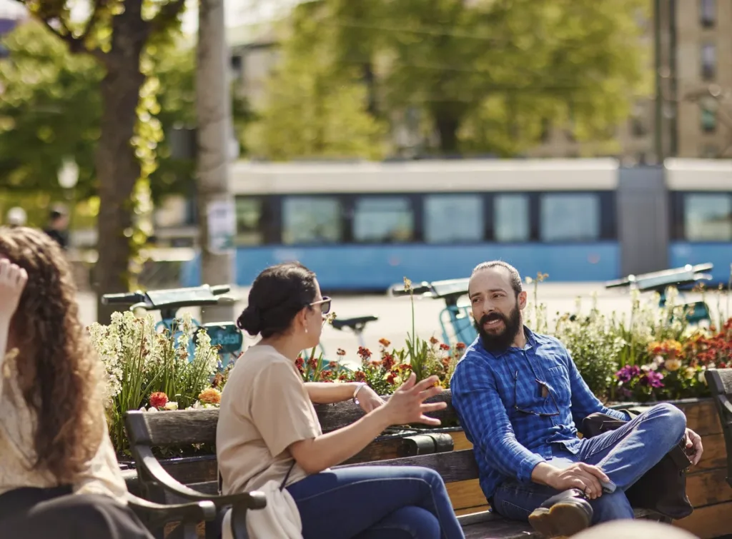 Två personer samtalar på en parkbänk omgiven av blommor; en spårvagn syns i bakgrunden.