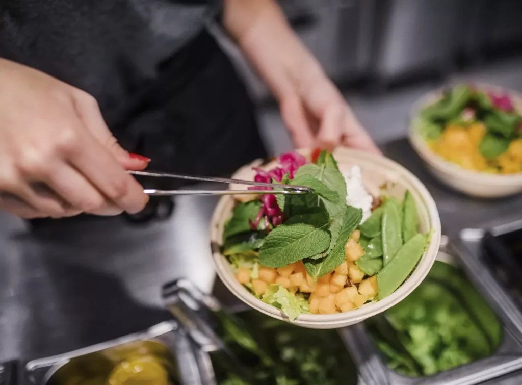 En hand håller tång och lägger gröna blad i en skål fylld med sallad, kikärtor och grönsaker.