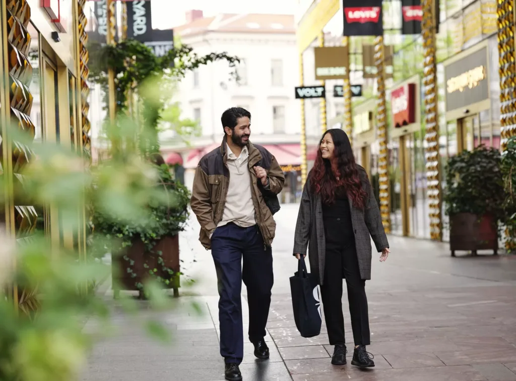 Ett par promenerar längs en shoppinggata med butiker och skyltar i bakgrunden.
