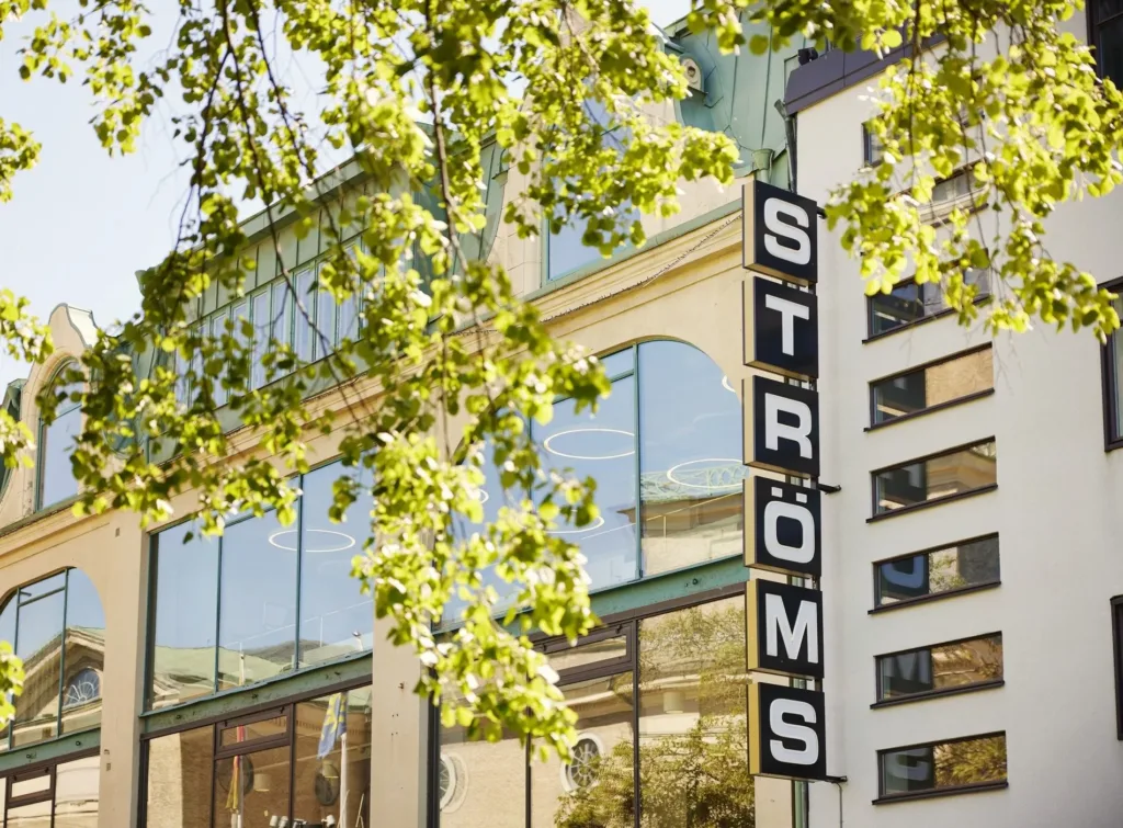 En byggnad med glasfönster och skylttexten "STRÖMS" synlig, omgiven av gröna trädgrenar.