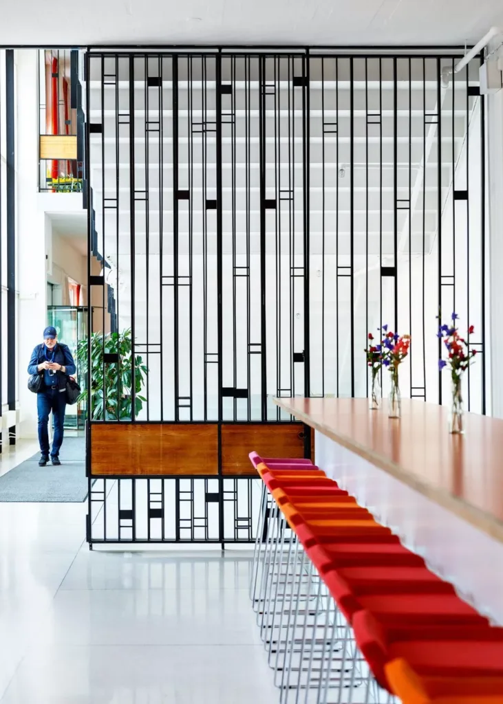 En modern foajé med en lång träbänk, röda stolar och en gallerdesign som delar av rummet. En person går genom en dörröppning i bakgrunden.