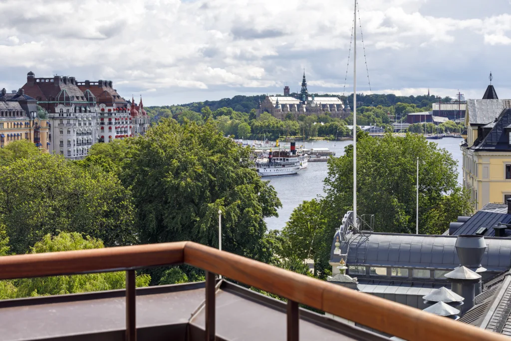 2. Utsikt över en flod med båtar, gröna träd och byggnader i bakgrunden från en balkong med trästaket.