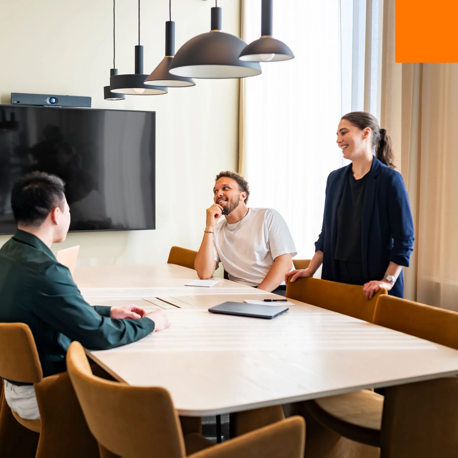 Tre personer sitter och står runt ett konferensbord i ett mötesrum med en TV på väggen.