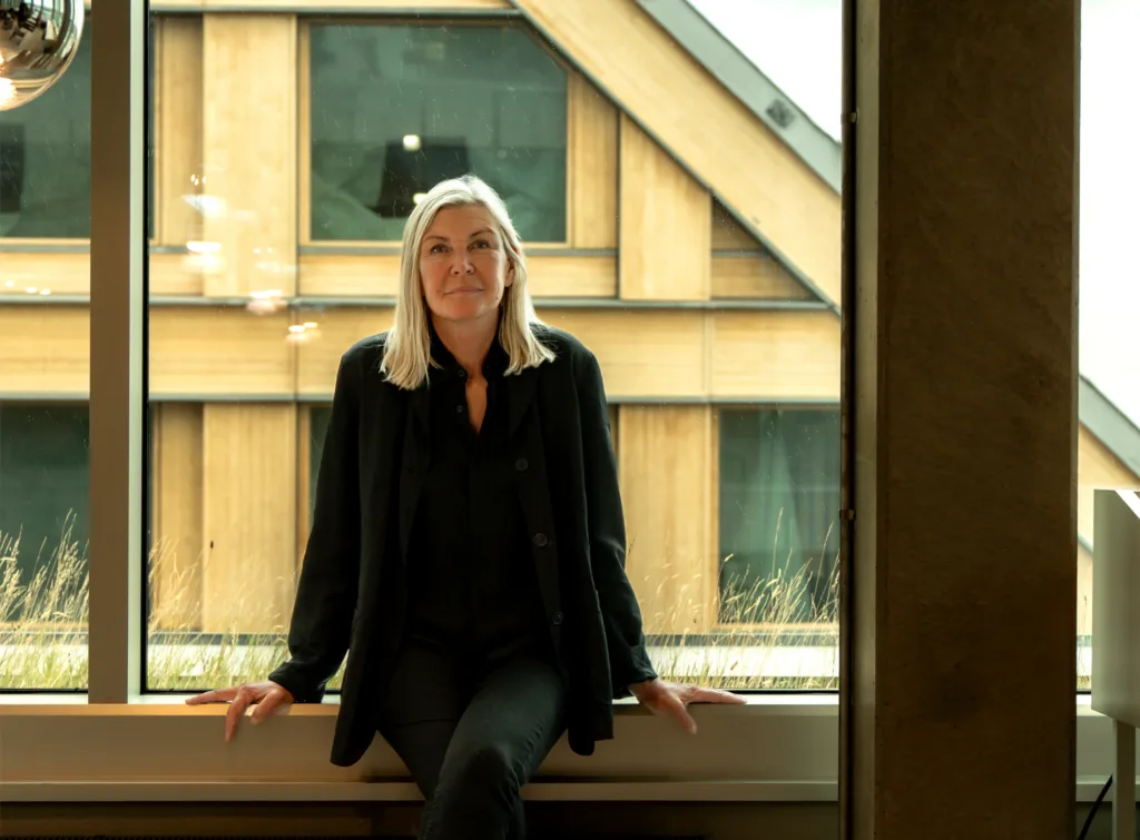 En kvinna med långt blont hår sitter avslappnat på en fönsterbräda, med en modern byggnad i trästil i bakgrunden.