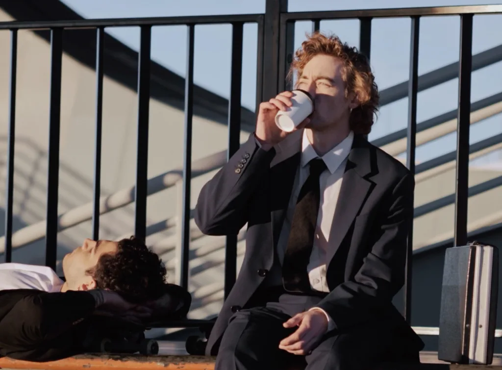 En person i kostym sitter utomhus och dricker ur en vit pappersmugg, medan en annan person ligger på en bänk i bakgrunden.