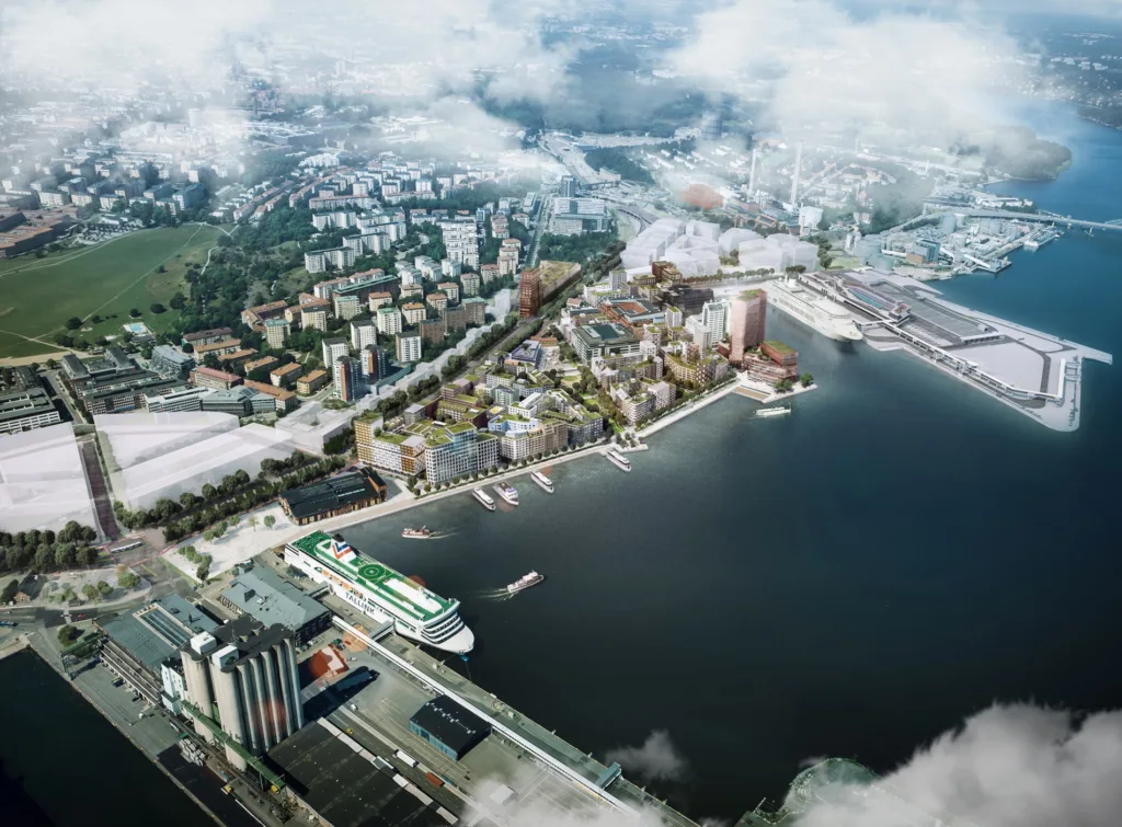 En vy över en stad vid vattnet med byggnader, båtar och grönområden, delvis täckt av moln.