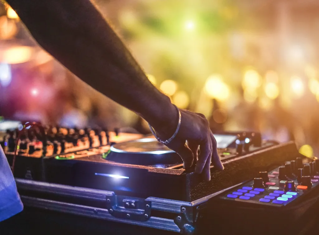 En DJ-mixer med en hand som justerar kontroller, med färgglada ljus och suddiga människor i bakgrunden.