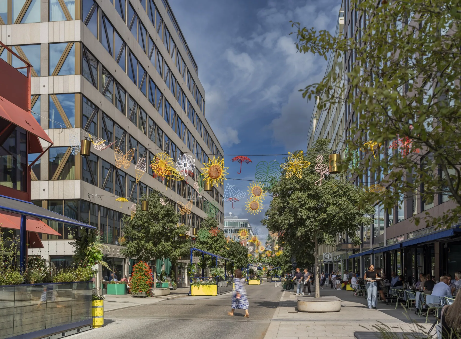 En stadsgata med höga byggnader, träd längs trottoaren och färgglada dekorationer hängande över vägen.