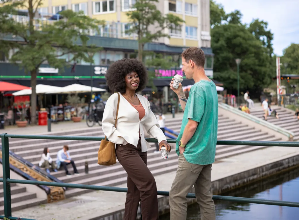 Två personer står och pratar vid en kanal med stadsmiljö och trappor i bakgrunden.