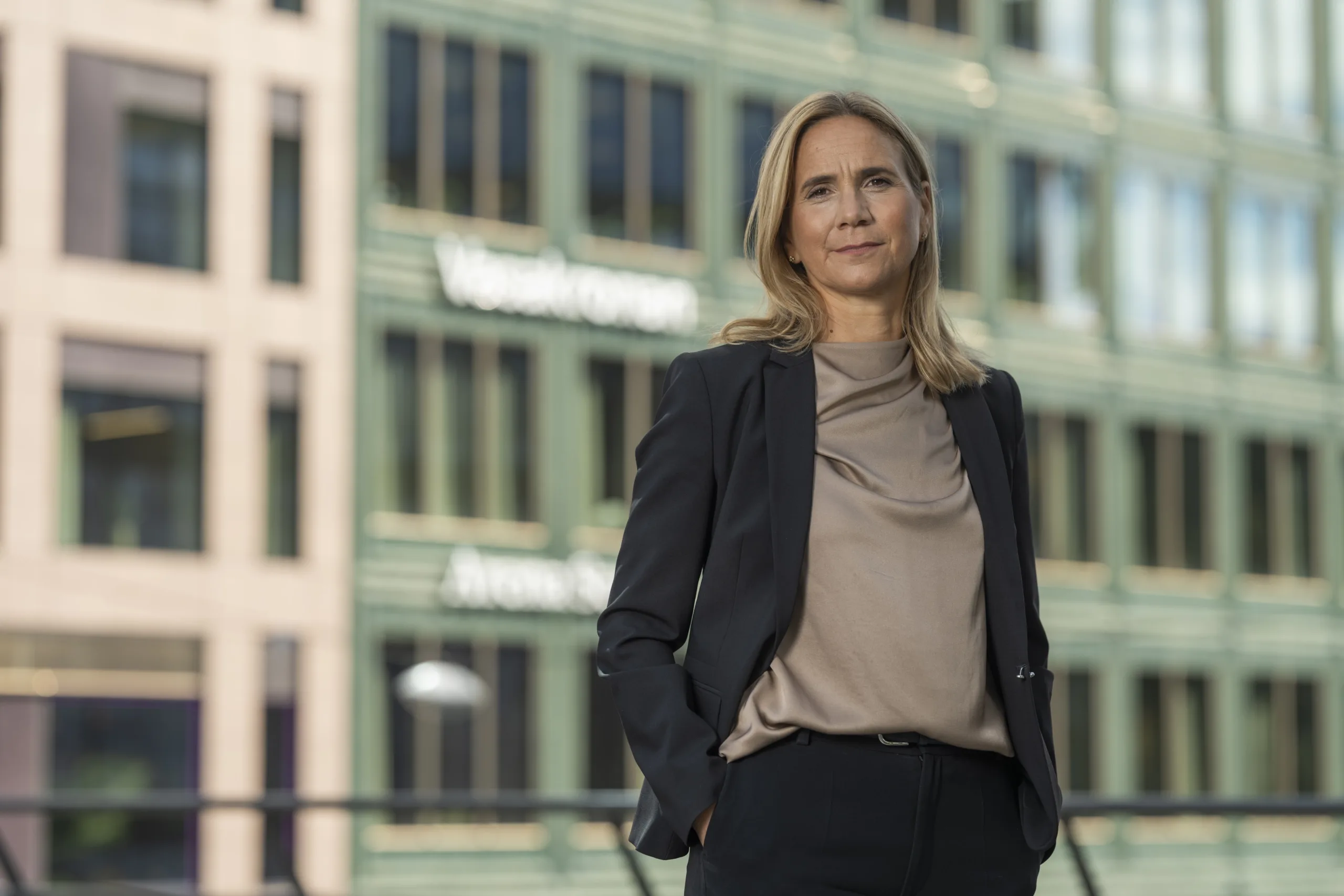 En kvinna i kostym står utomhus med händerna i fickorna, framför en byggnad med texten "Vasakronan" synlig på fasaden.